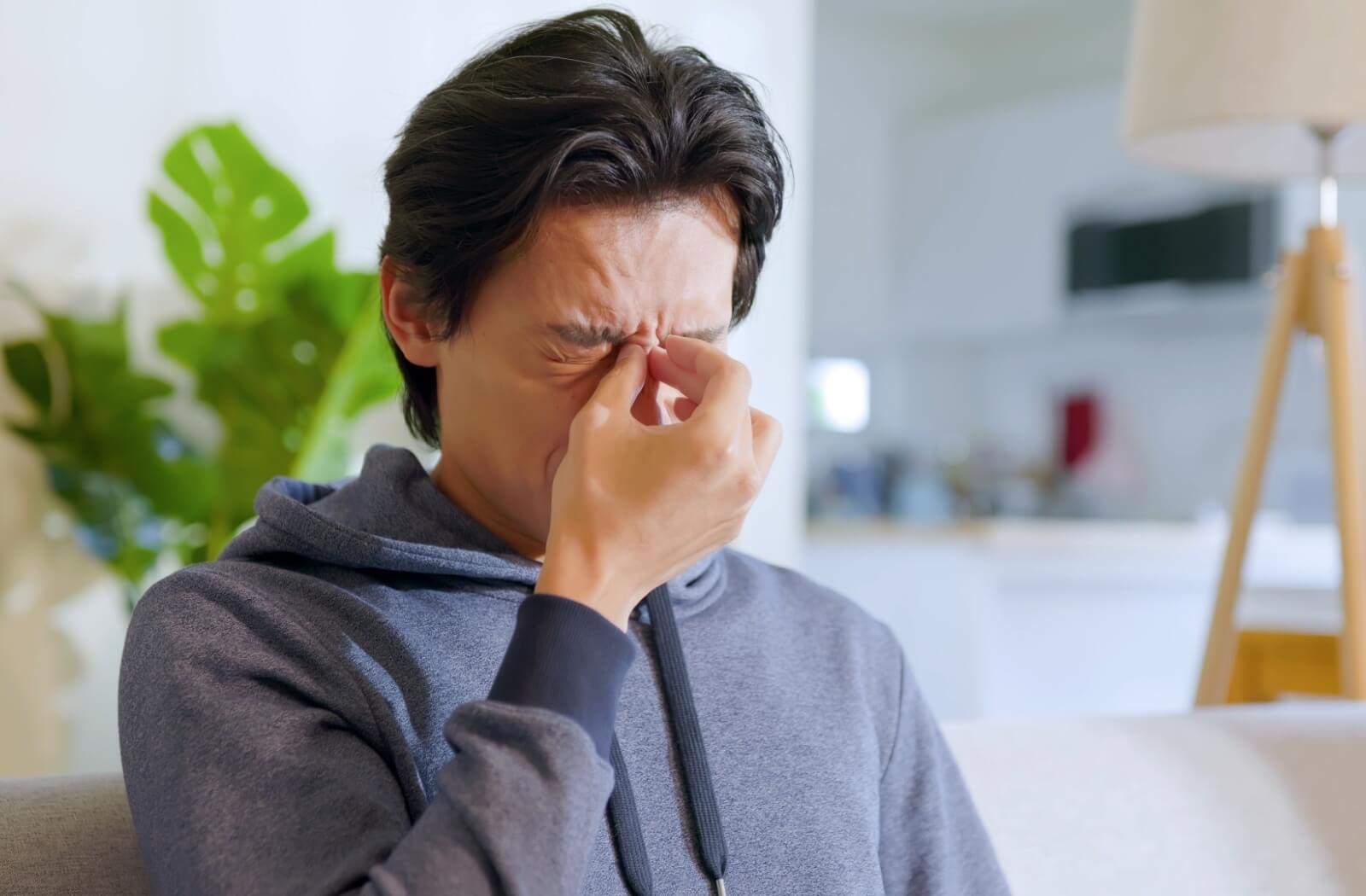 Person sitting on a couch wearing a blue hoodie, pinching the bridge of their nose with eyes closed, appearing to experience eye strain or discomfort.