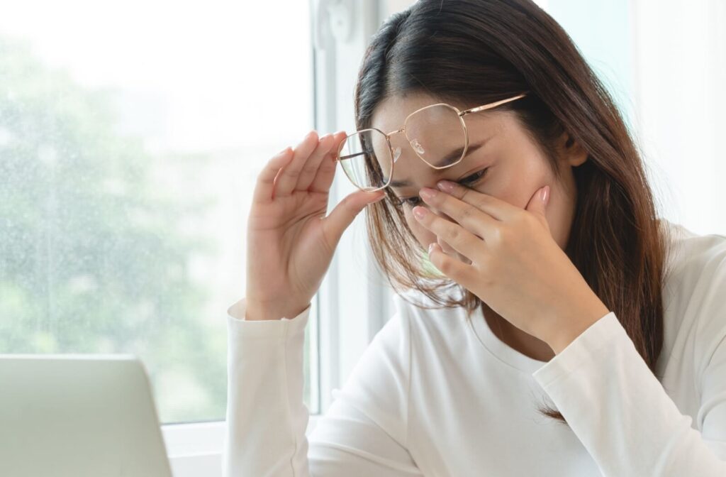 A person in a white top lifting their gold-rimmed glasses and rubbing their eyes while sitting at a desk with a laptop, illustrating symptoms of digital eye strain and dry eye discomfort.